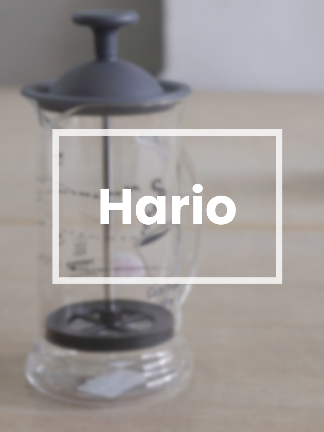 Glass coffee press with black lid and base on a wooden surface, featuring the brand 'Hario'.